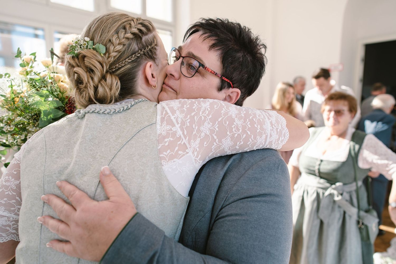 Two people hugging and showing affection in a bright indoor setting with guests in the background.