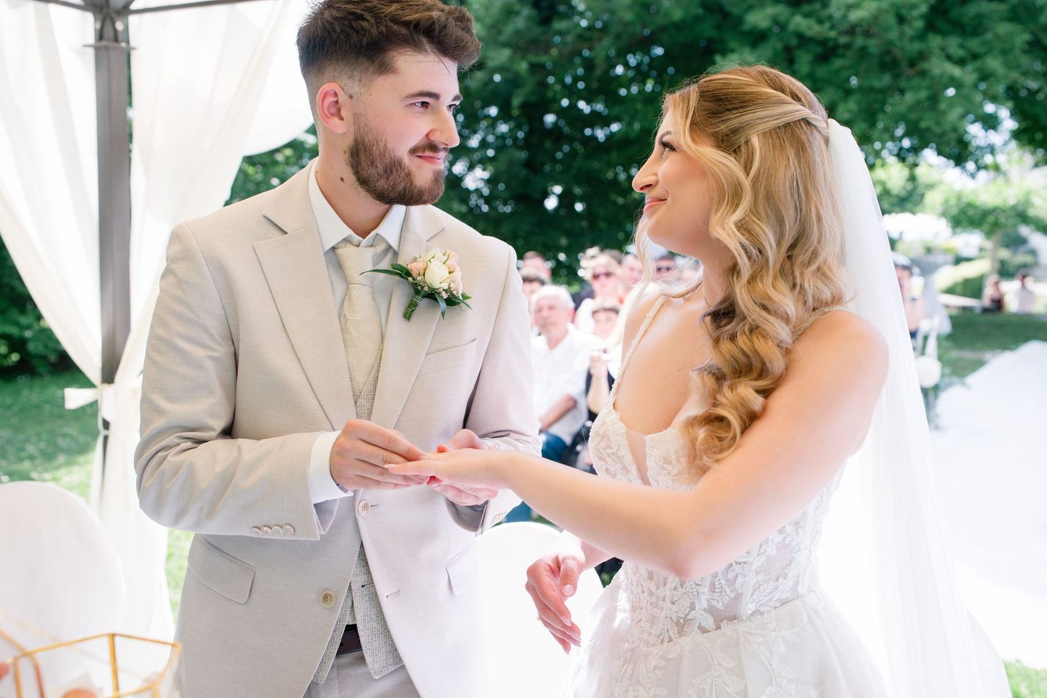 Bride and groom exchanging rings outdoors at a wedding ceremony, smiling joyfully in elegant attire.