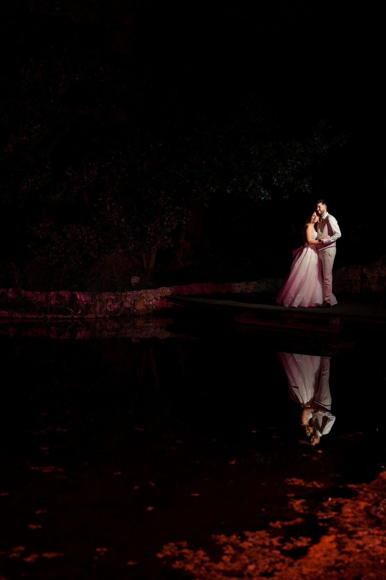 A bride and groom embrace near a reflective pond at night, illuminated by warm romantic lighting.
