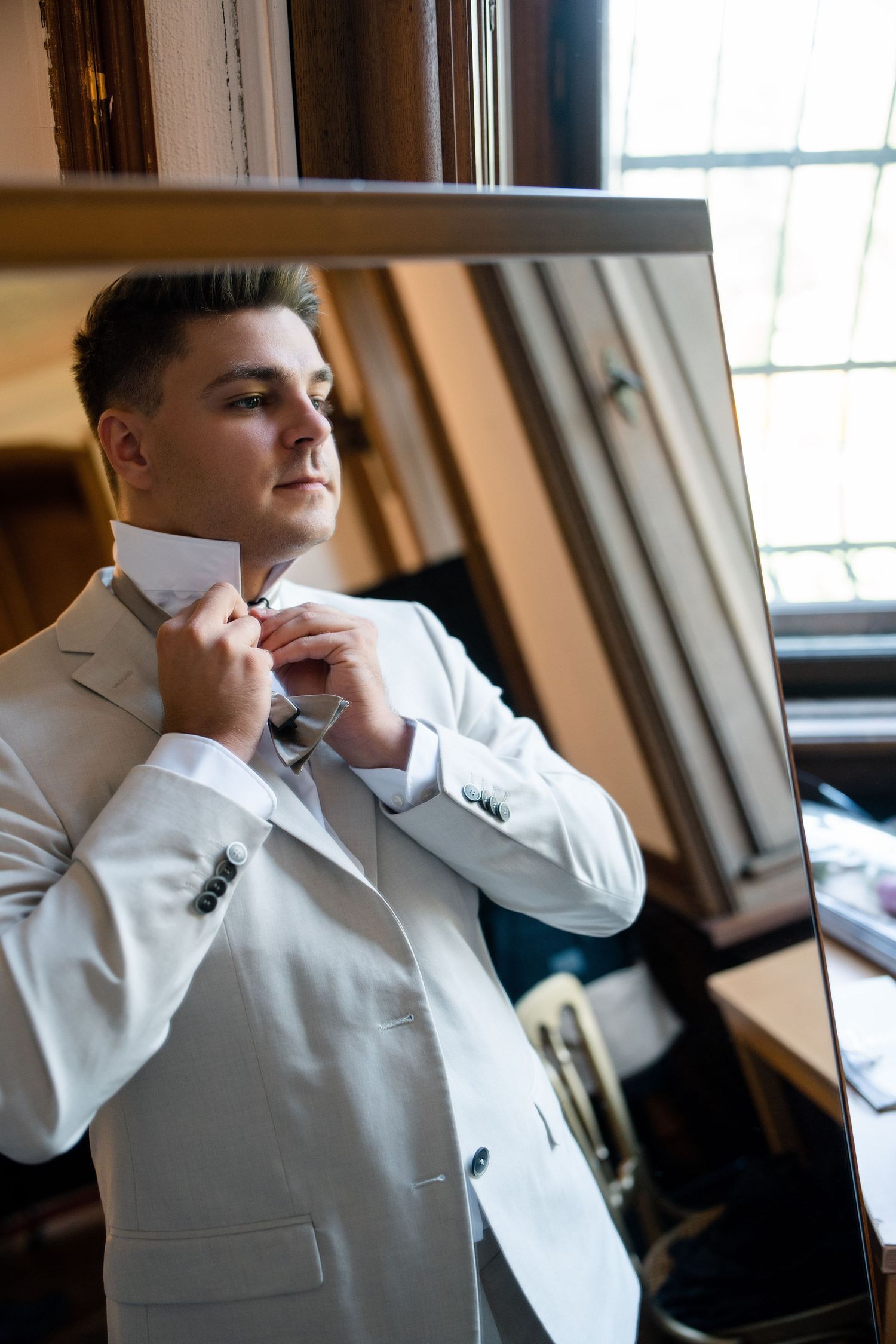 Person in white suit adjusts bowtie while looking in mirror during formal preparation.