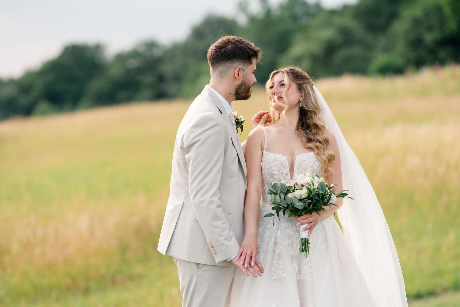 A bride and groom holding hands in a sunlit field, the bride wearing a white gown with veil and holding a bouquet.