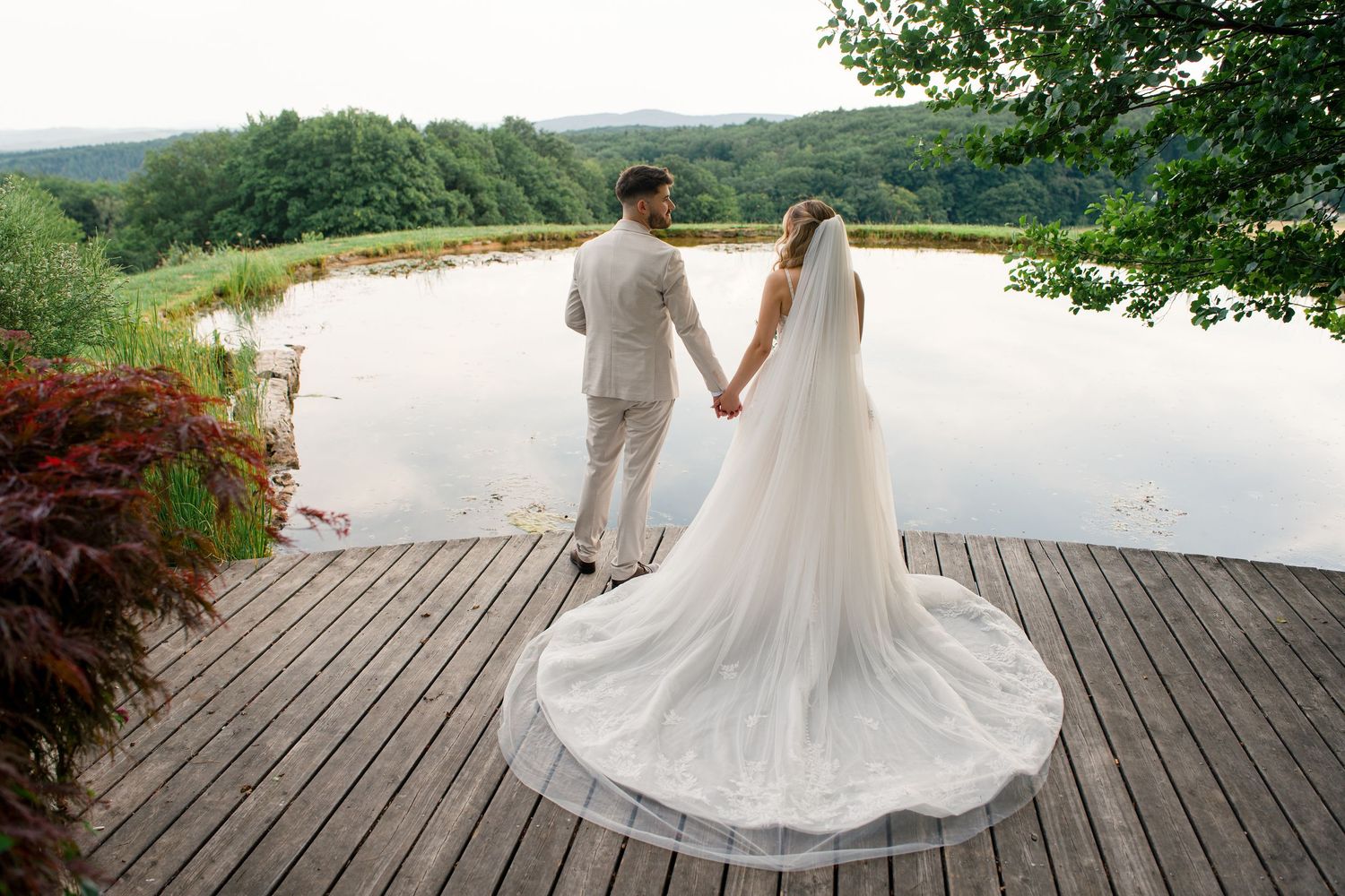Bride and groom holding hands on wooden deck overlooking serene lake surrounded by lush green forest.