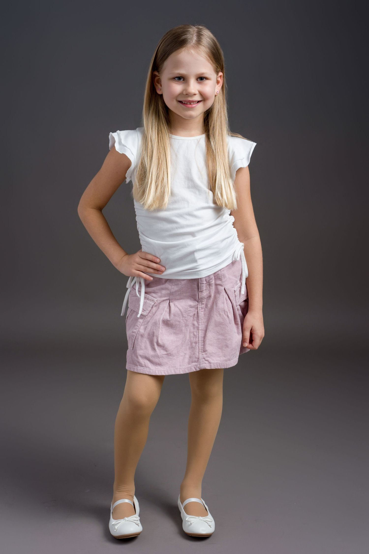 Child wearing white top and pink skirt poses confidently with hand on hip against gray background.