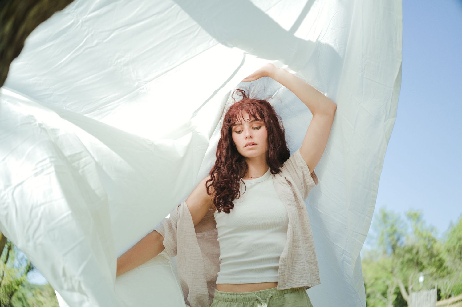 Young woman with curly auburn hair posing outdoors with flowing white fabric billowing around her in sunlight.