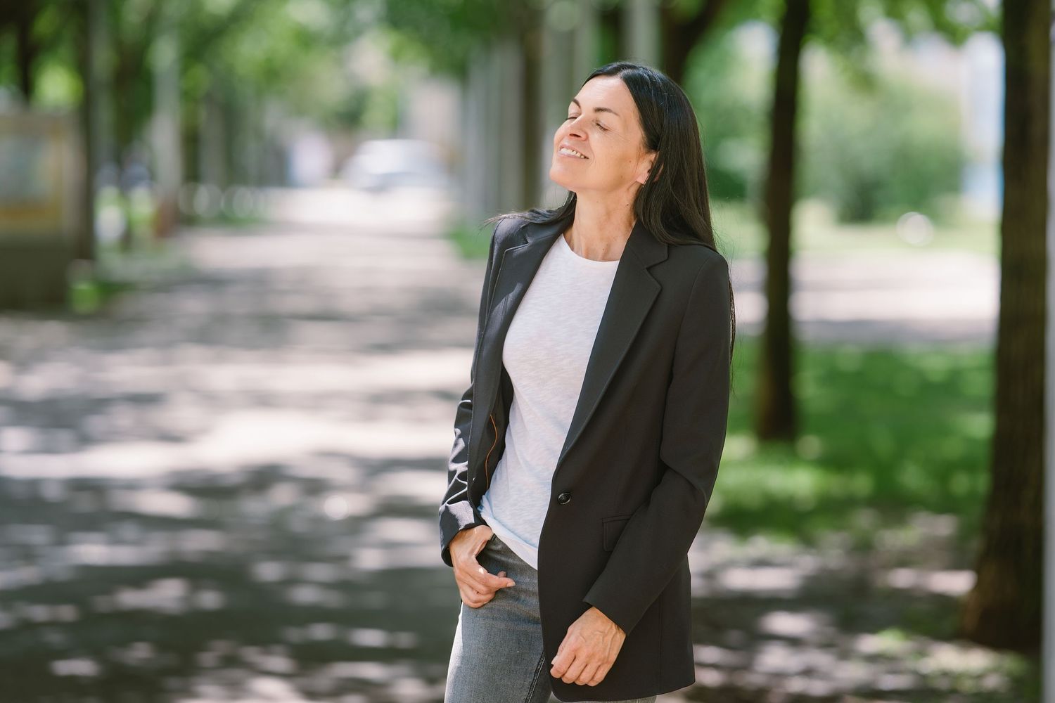 Woman in black blazer and jeans enjoying fresh air outdoors in a tree-lined park on a sunny day.
