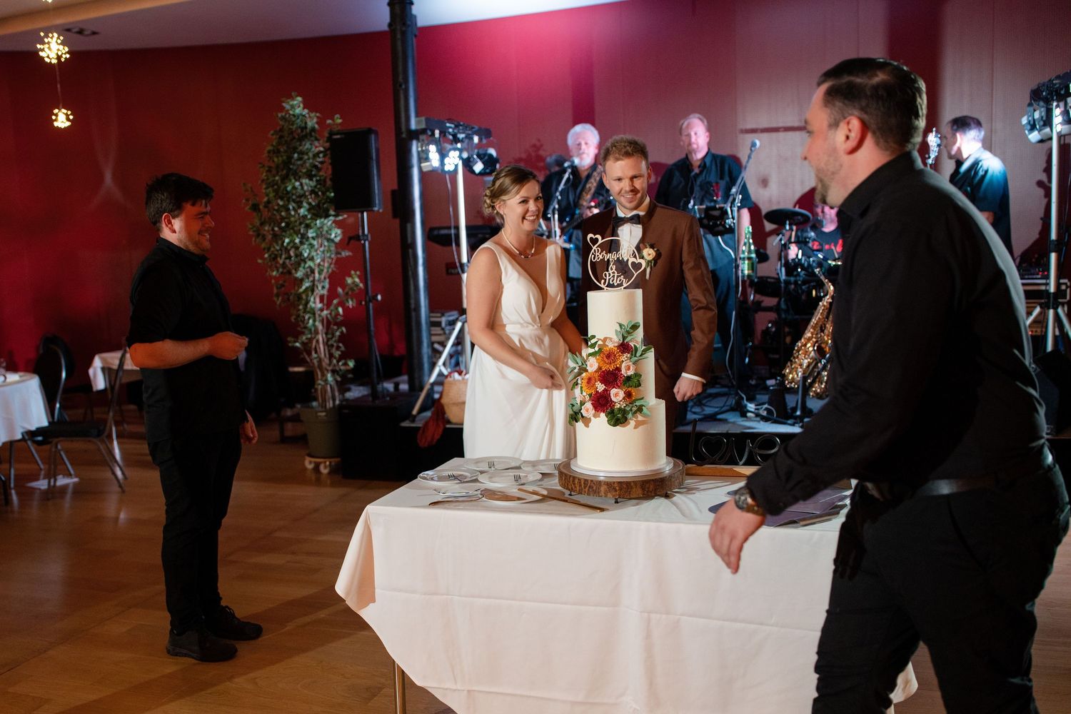 Wedding cake cutting ceremony in a reception venue with red walls and stage lighting.