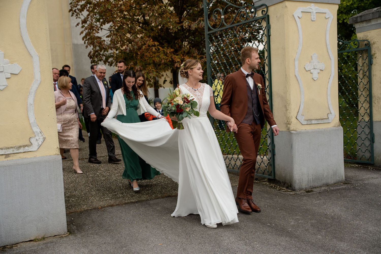 Wedding party exits a yellow building with decorative architectural details during celebration.