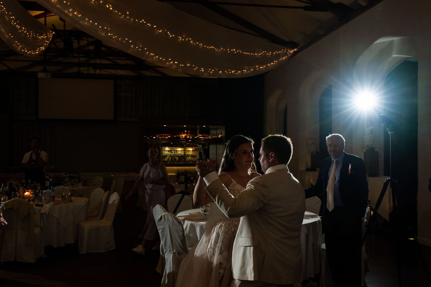 Couple shares first dance at elegant indoor wedding reception with string lights and soft lighting.