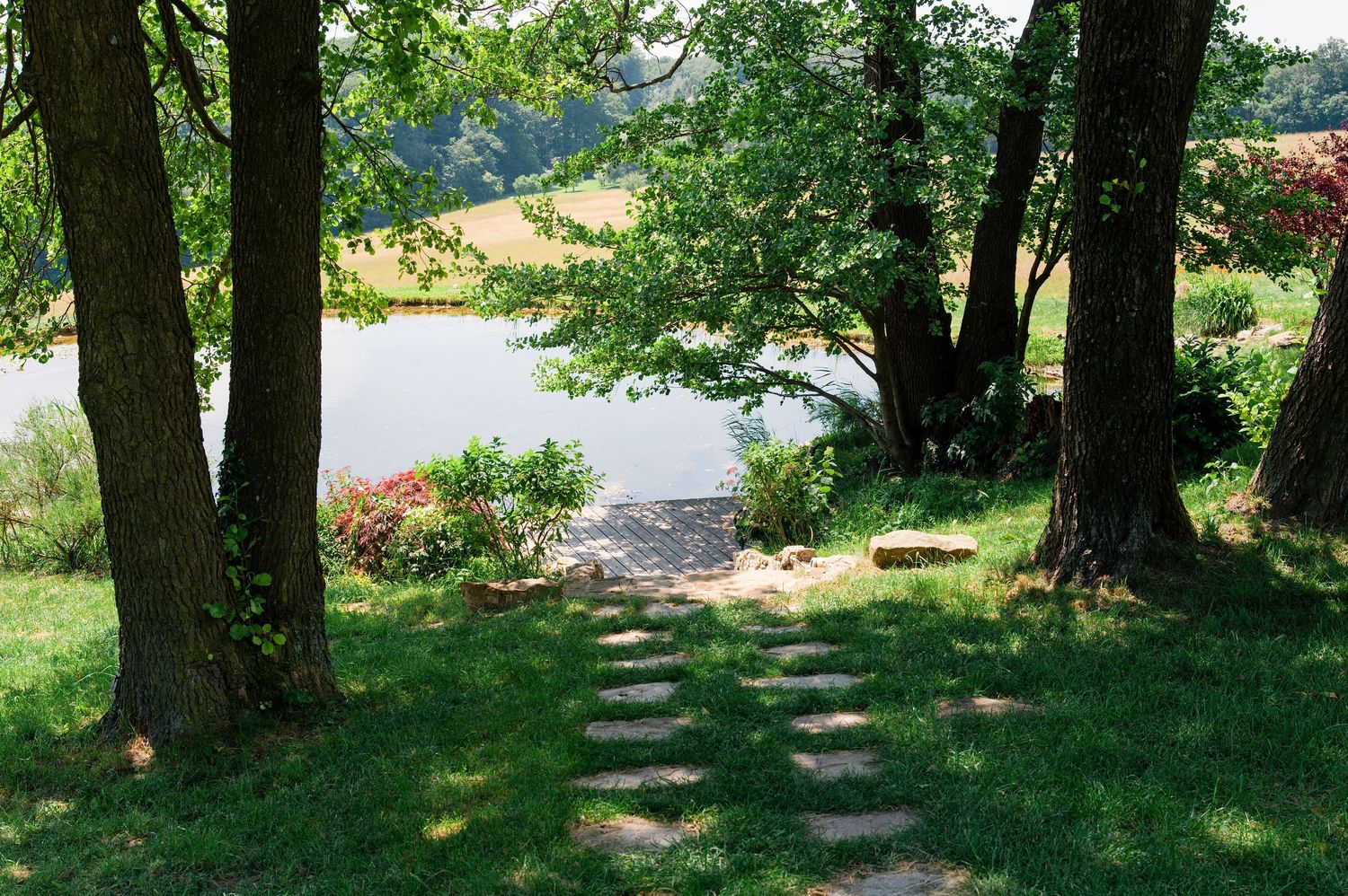 Stone path leads through shaded trees to a serene lakeside dock surrounded by lush green grass.