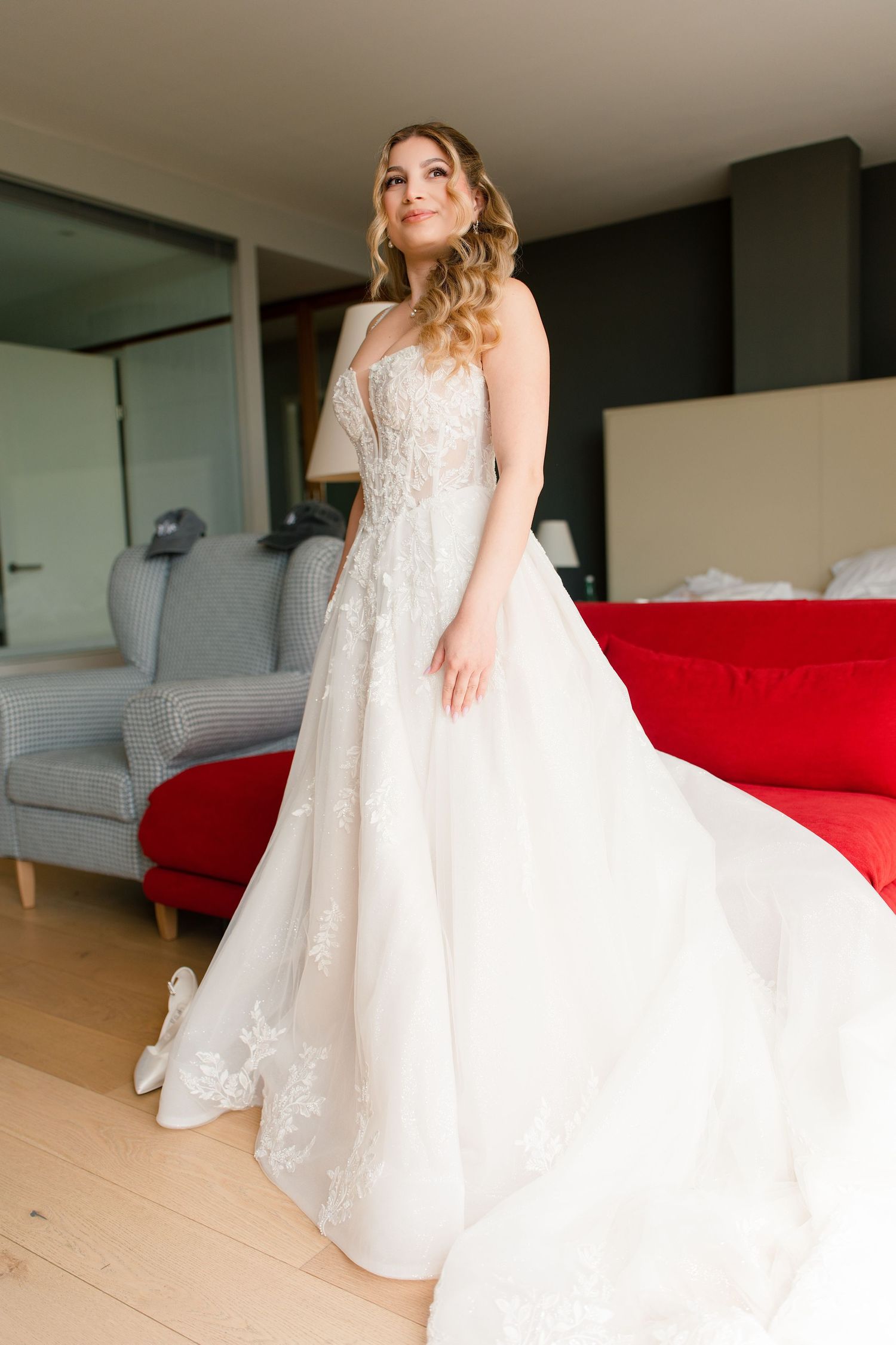 Bride in strapless lace wedding gown with flowing skirt posing in hotel room with red sofa.