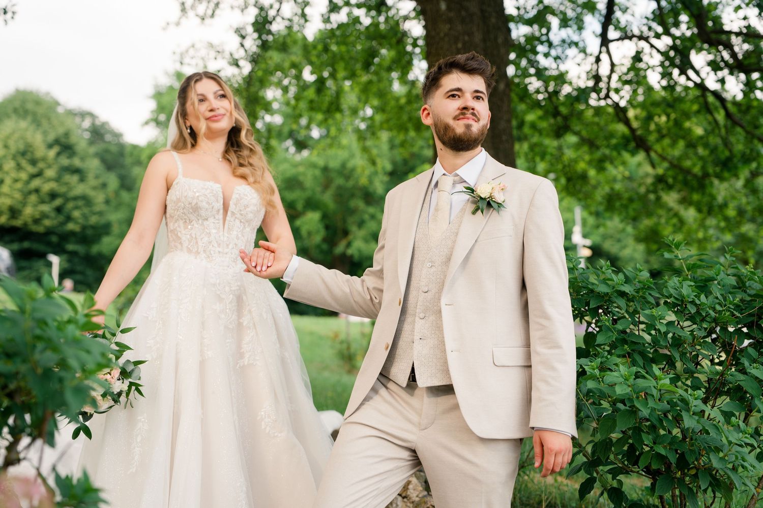 Bride in strapless lace gown holds hands with groom in tan suit with floral boutonniere outdoors.