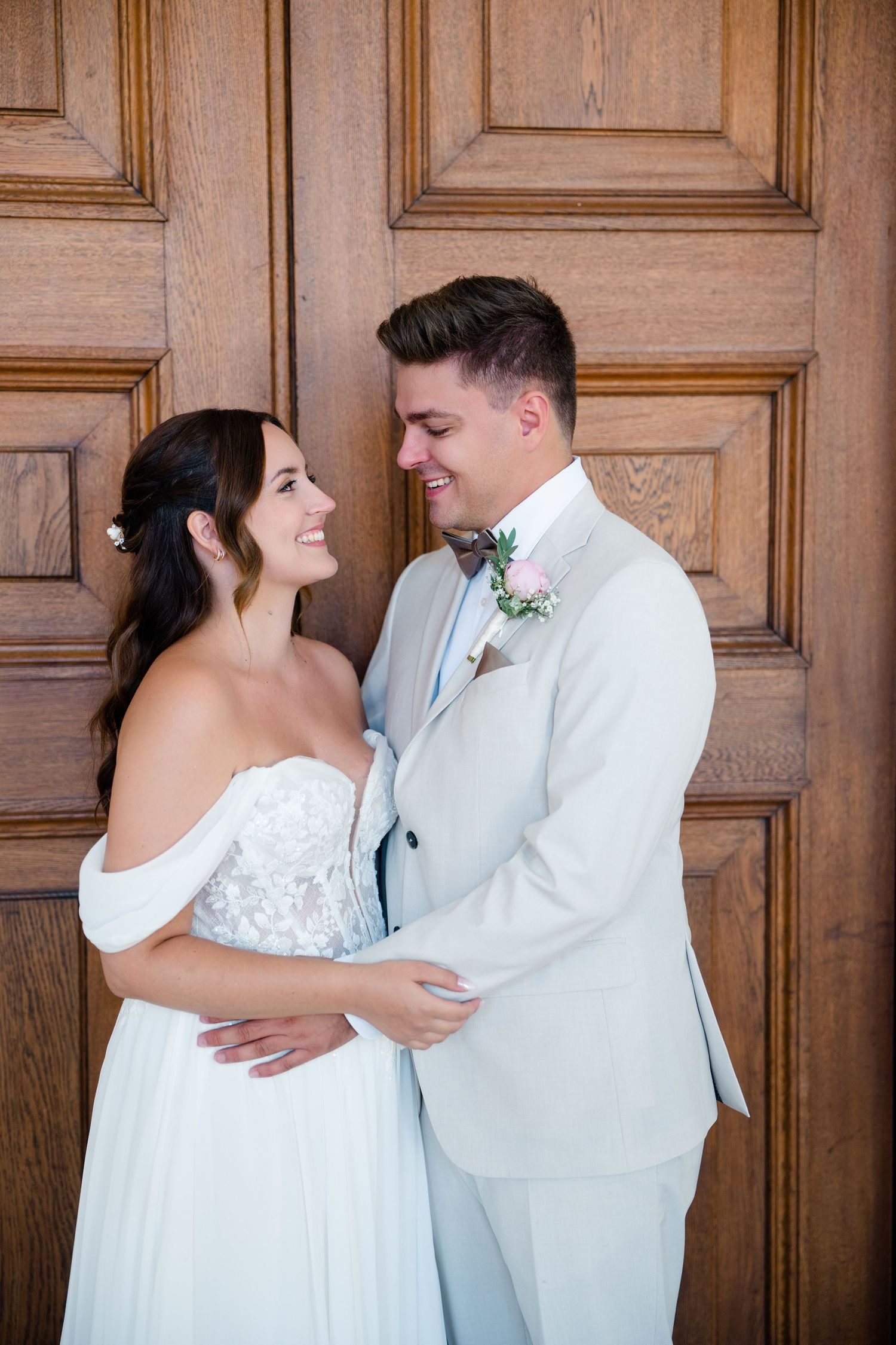 Newlyweds in white attire share an intimate moment against a wooden door backdrop.