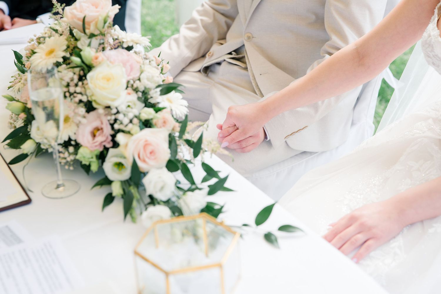 Bride and groom holding hands at signing table with elegant white and pink floral arrangement at wedding ceremony.