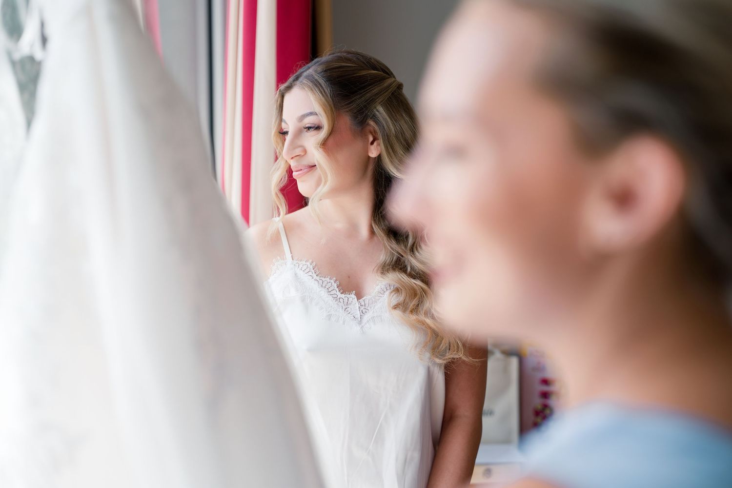 Smiling bride in white lace dress standing by window with wedding gown hanging nearby during bridal preparations.