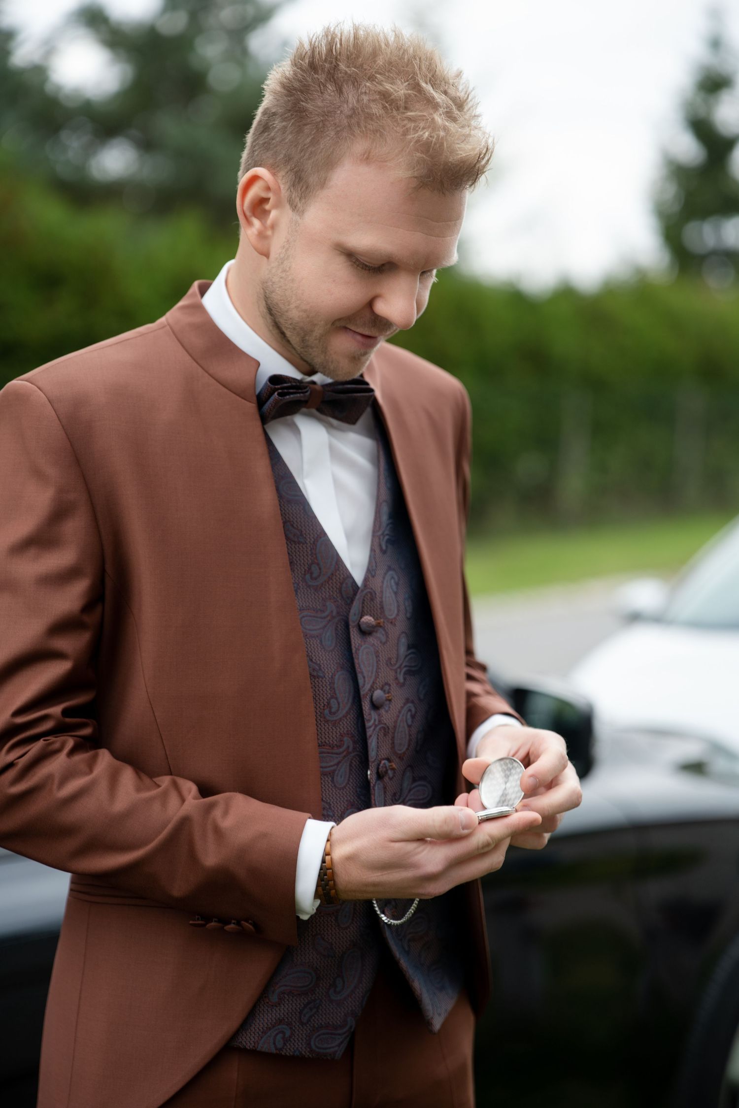 Groom in brown suit and bowtie checks his pocket watch outdoors before the ceremony.