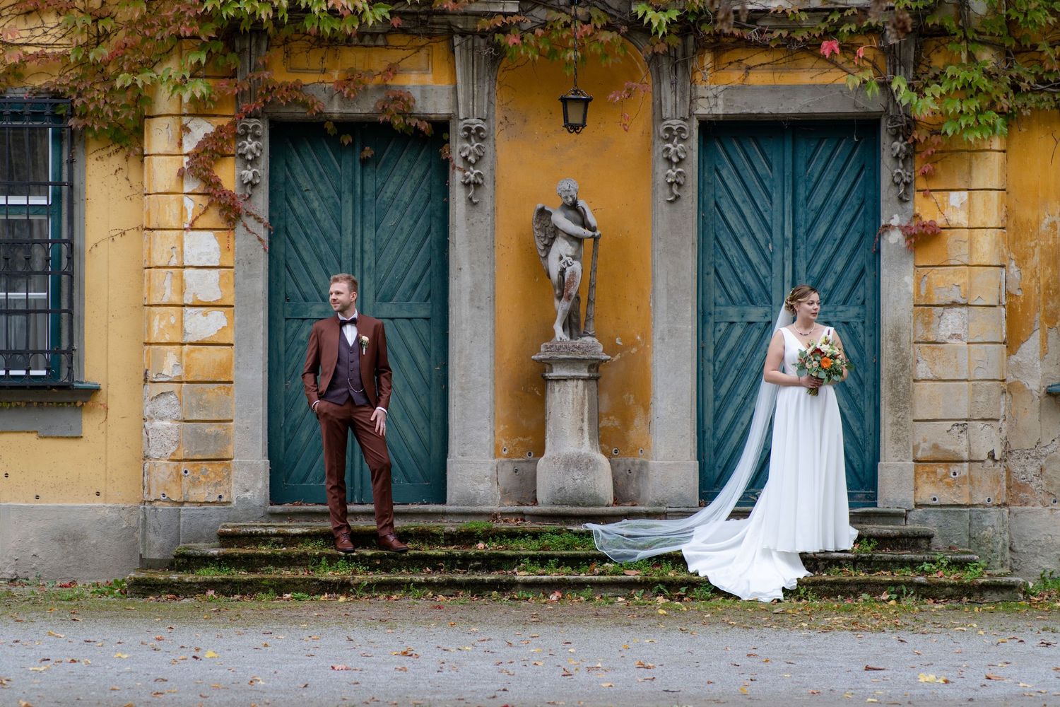 Couple standing apart by teal doors and statue against yellow building exterior.