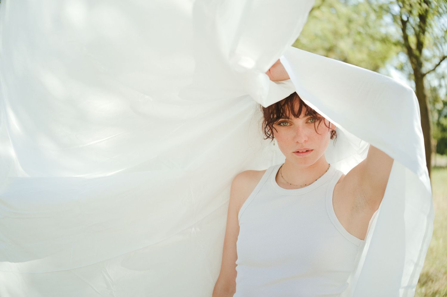 A young woman in a white tank top holds flowing white fabric overhead outdoors on a sunny day.