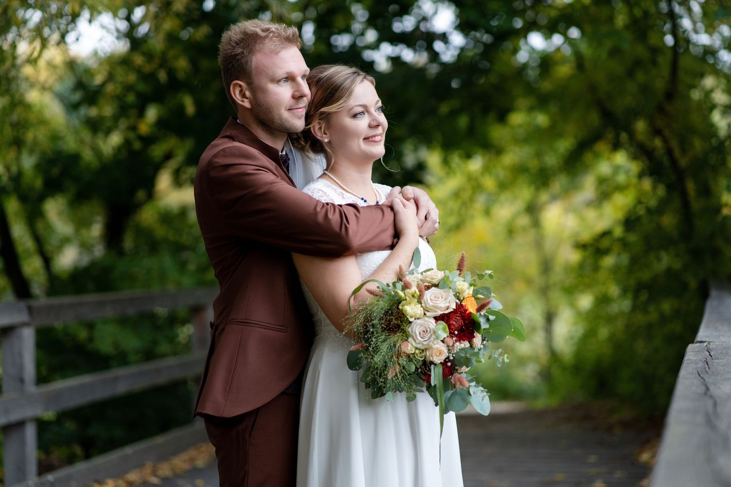 Couple in brown suit and white dress embrace while holding floral bouquet outdoors.