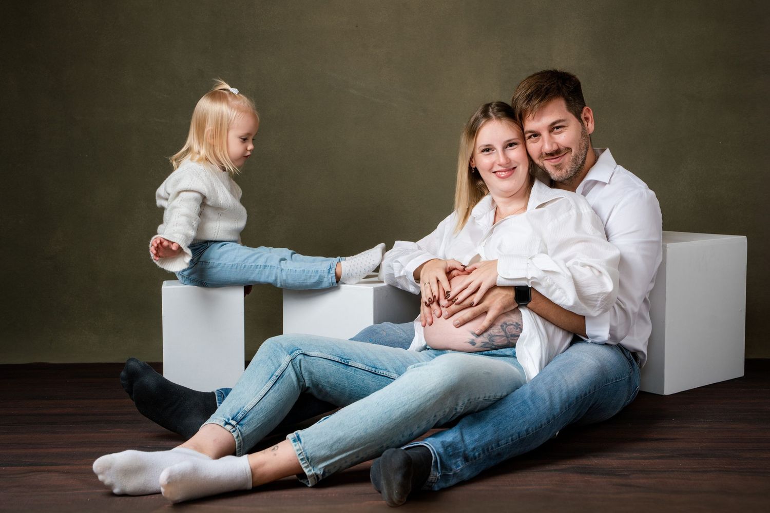 A family in casual white and denim outfits sits together on white blocks against a dark green studio backdrop.