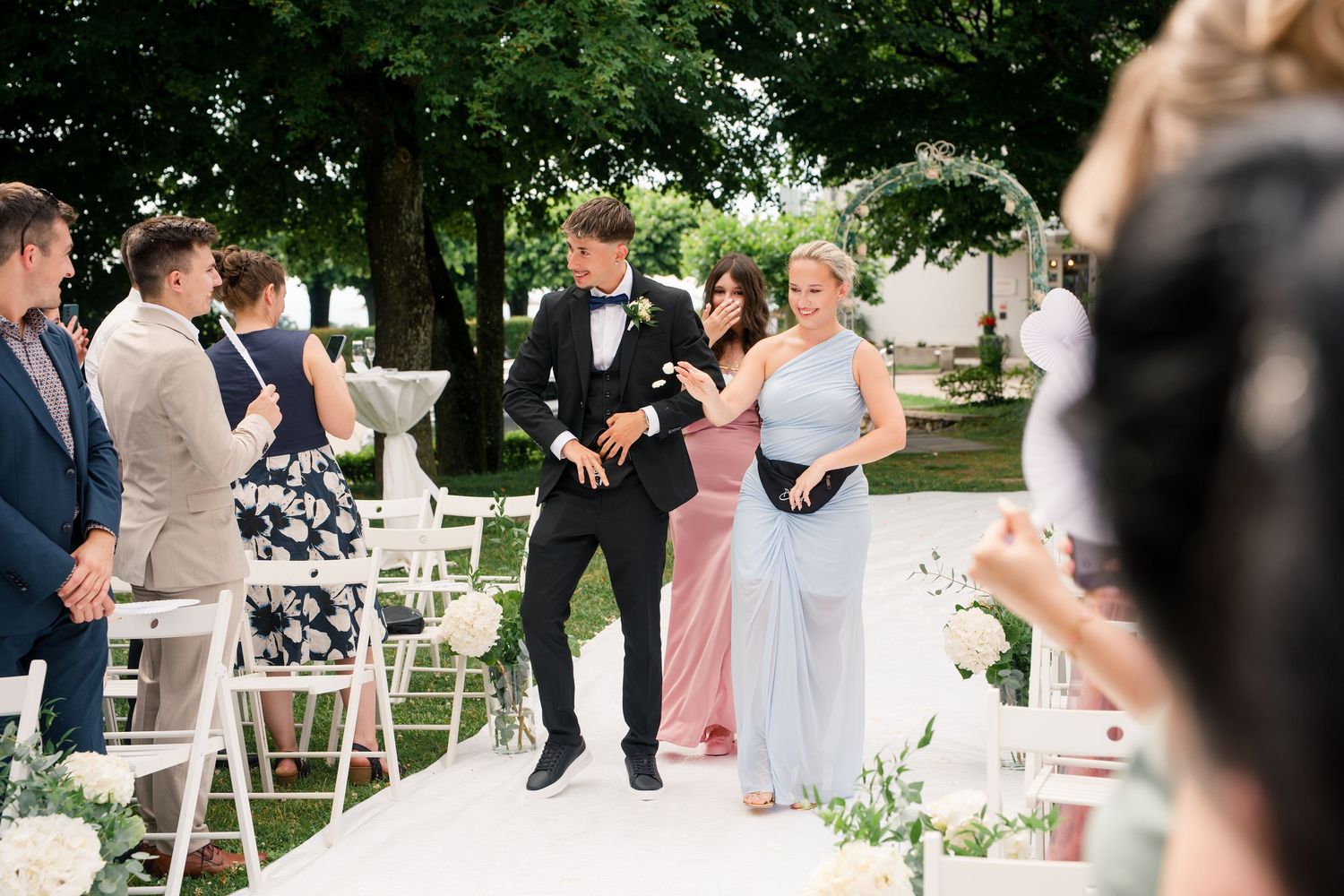 Groom in tuxedo walks down wedding aisle with bridesmaid in blue dress at elegant outdoor ceremony.