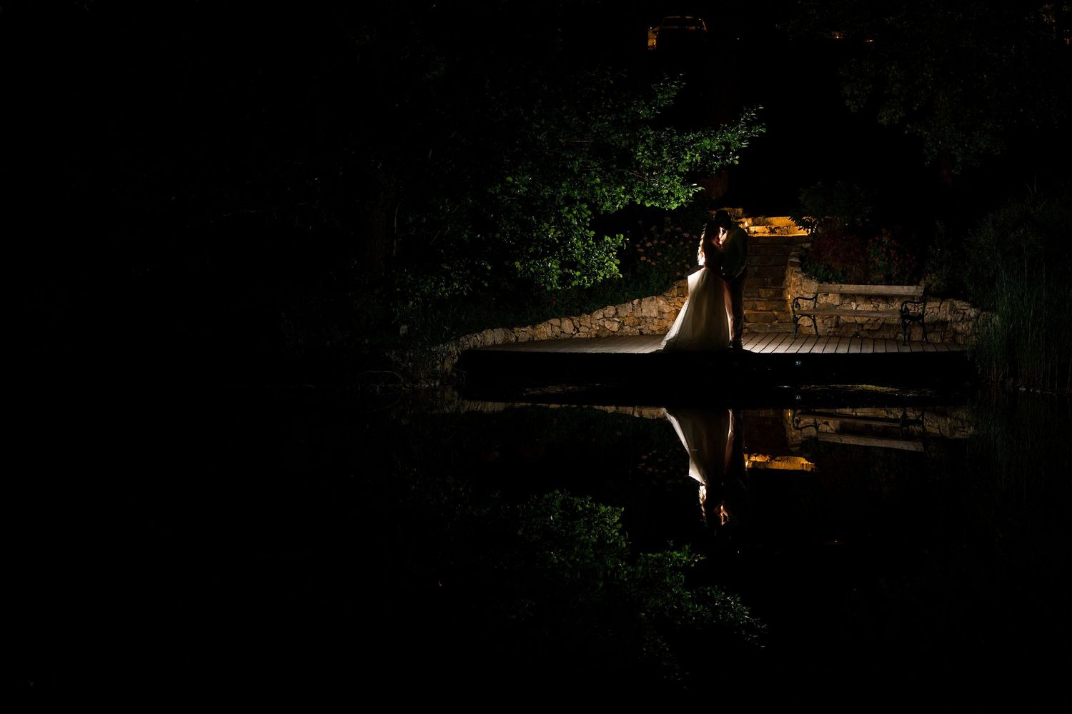 Nighttime reflection of an illuminated bridge over still dark water surrounded by lush green trees.