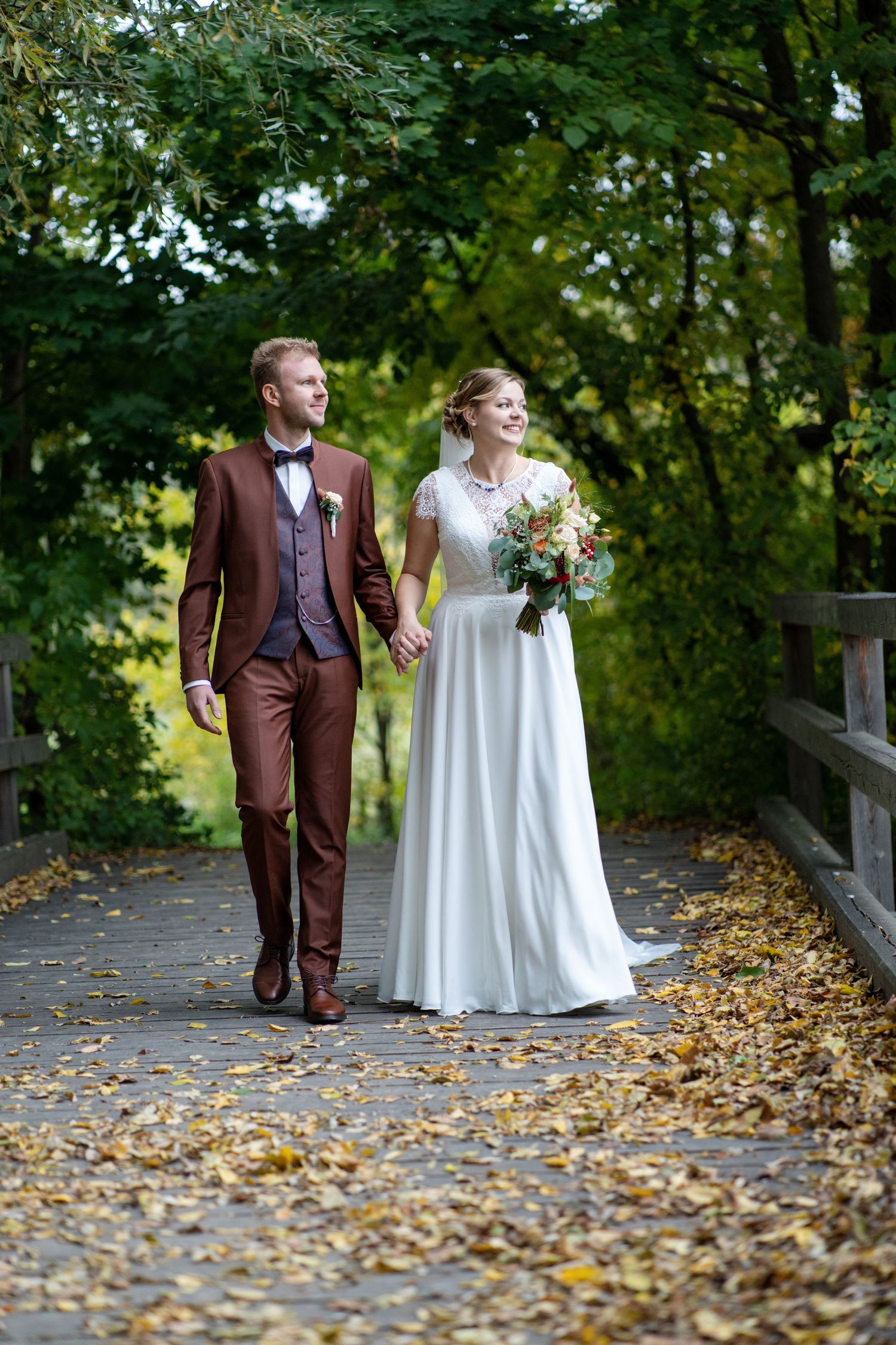 A couple in wedding attire walking along a leaf-covered path surrounded by autumn trees.