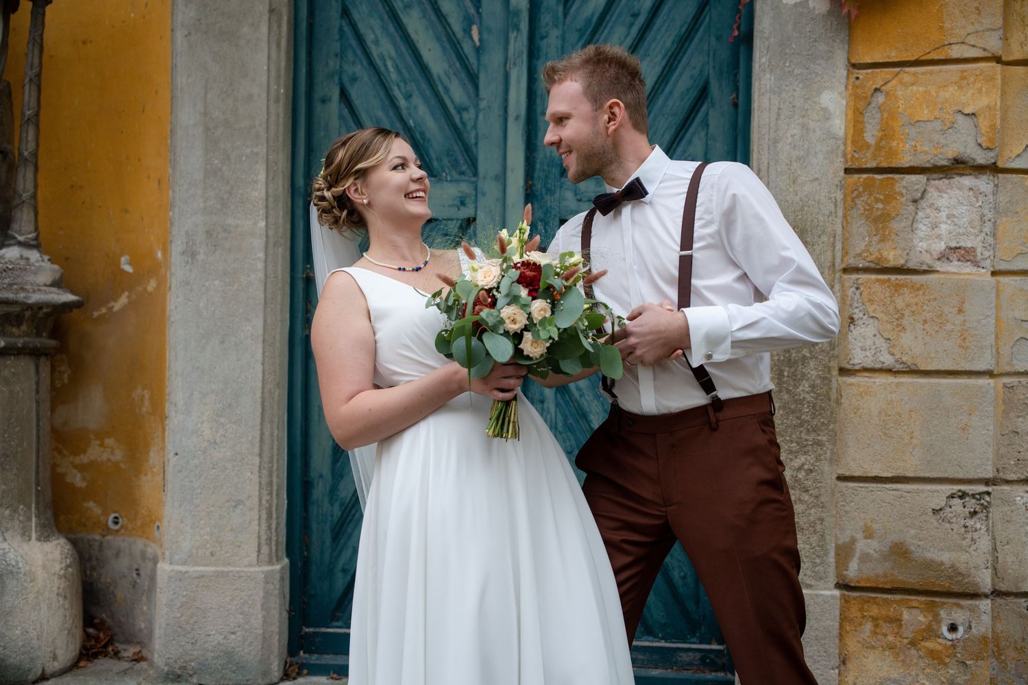 Newlyweds pose against rustic yellow stone walls and teal doors for romantic wedding portraits.