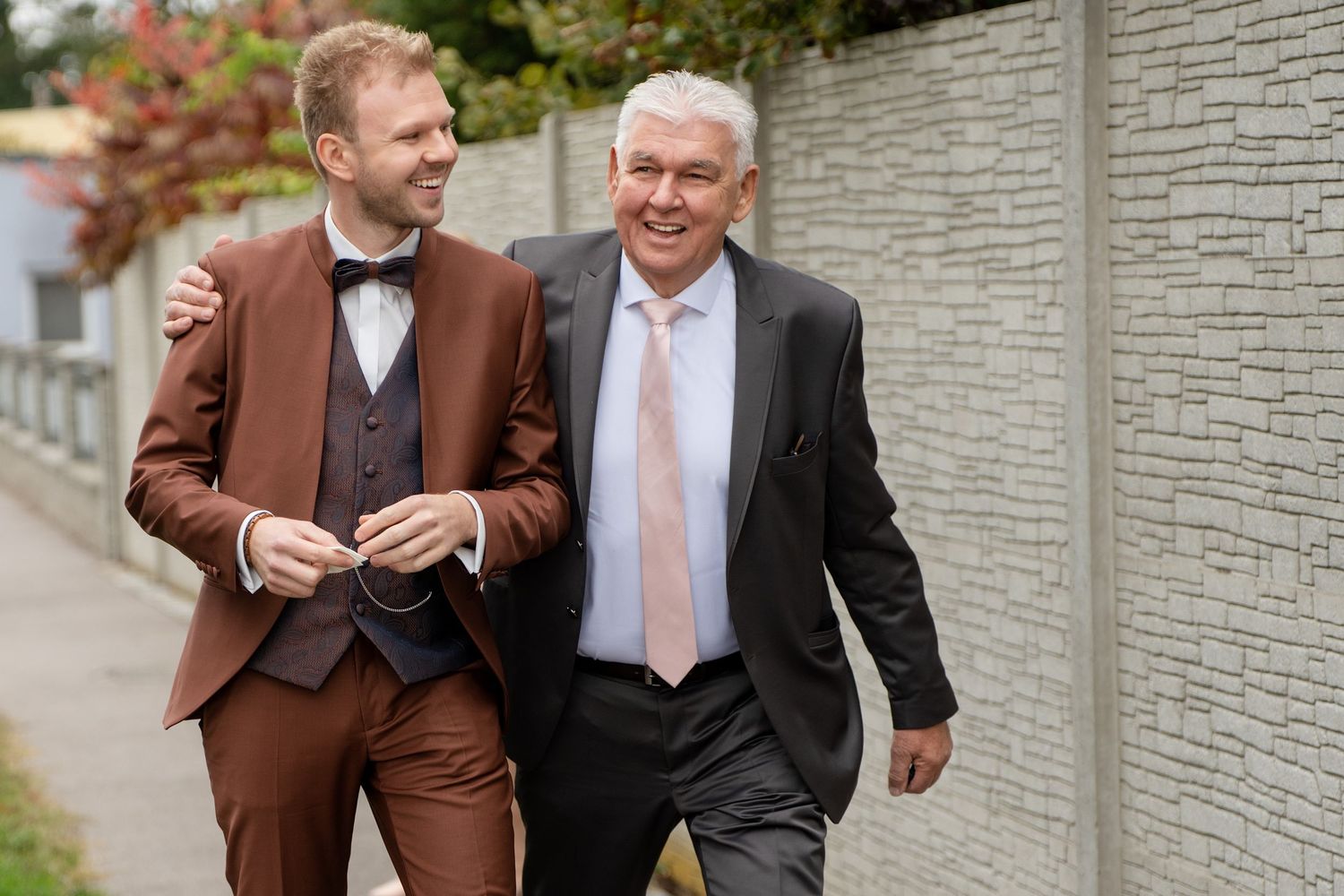 Two people in formal attire walk together and smile while wearing suits along a gray wall outdoors.