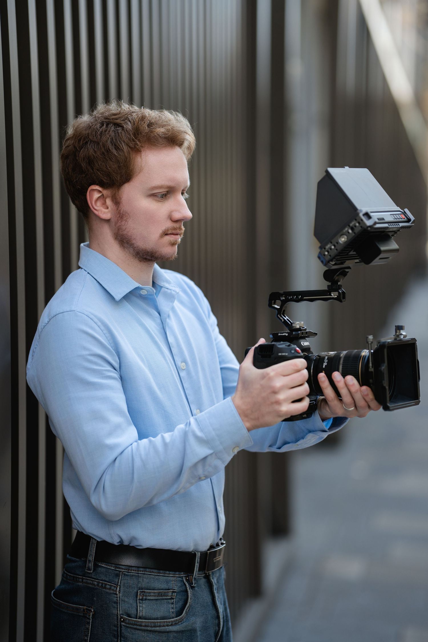 Photographer in light blue shirt and jeans demonstrates various camera holding positions against a striped wall background.