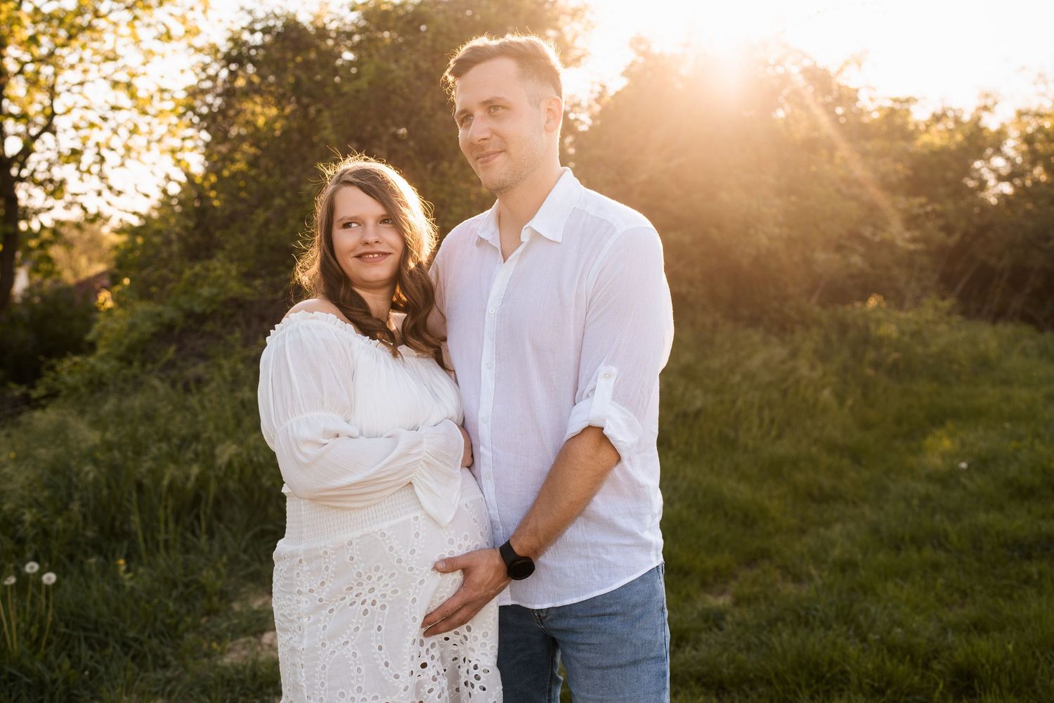 A couple embraces in a sunlit outdoor setting with golden light filtering through trees in the background.