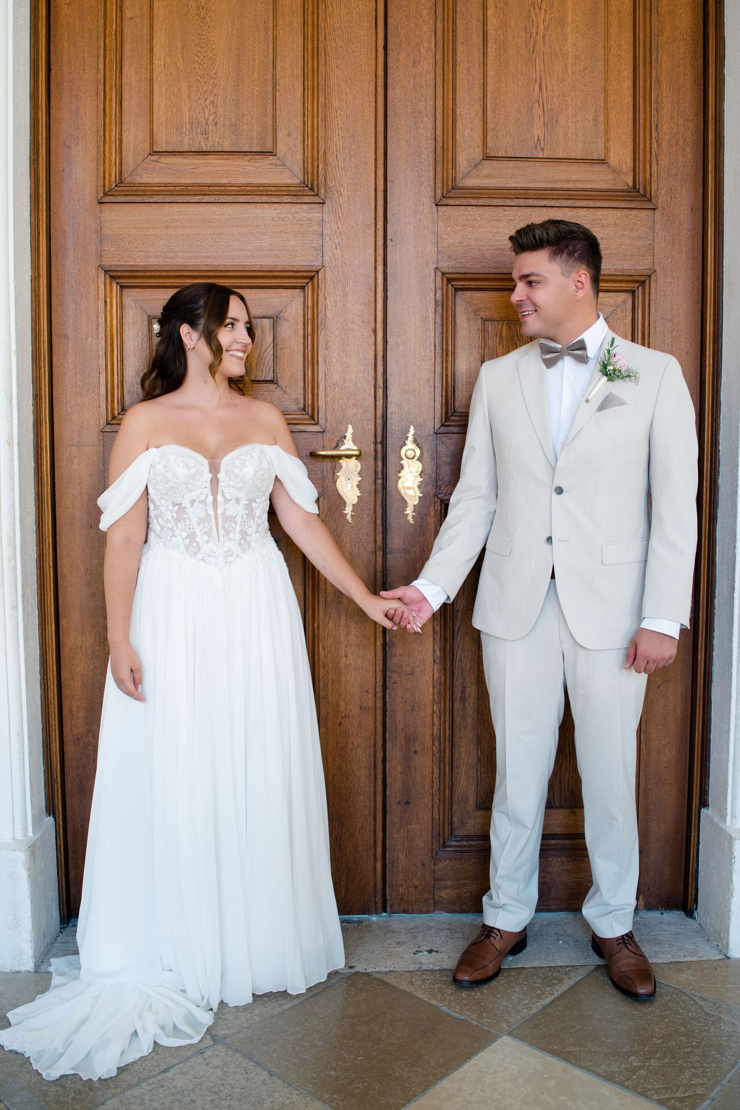 Wedding couple poses by grand wooden doors, bride in flowing dress and groom in light suit.