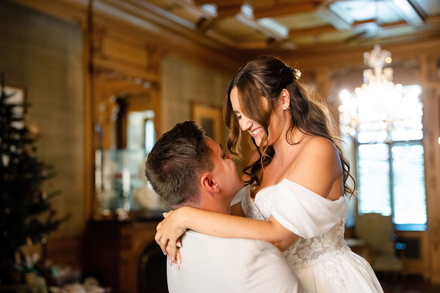 Wedding couple dancing in a grand ballroom with wooden paneled walls and chandelier.