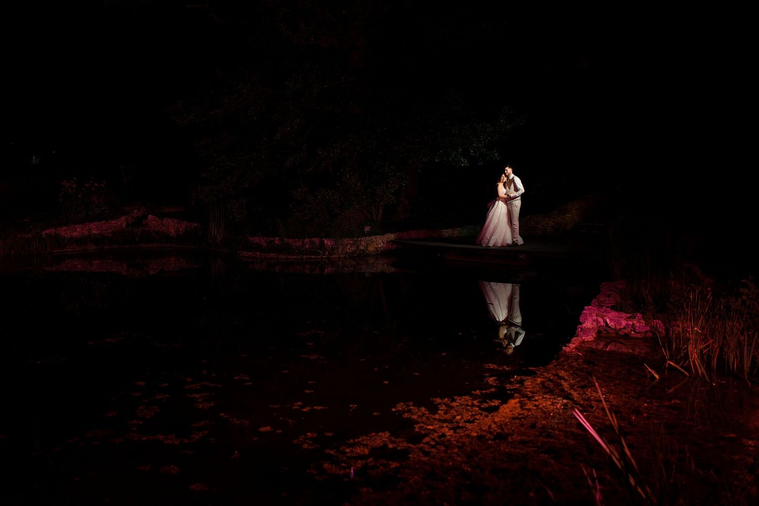 Couple embracing by a reflective pond at night, illuminated by dramatic red lighting in a romantic outdoor setting.