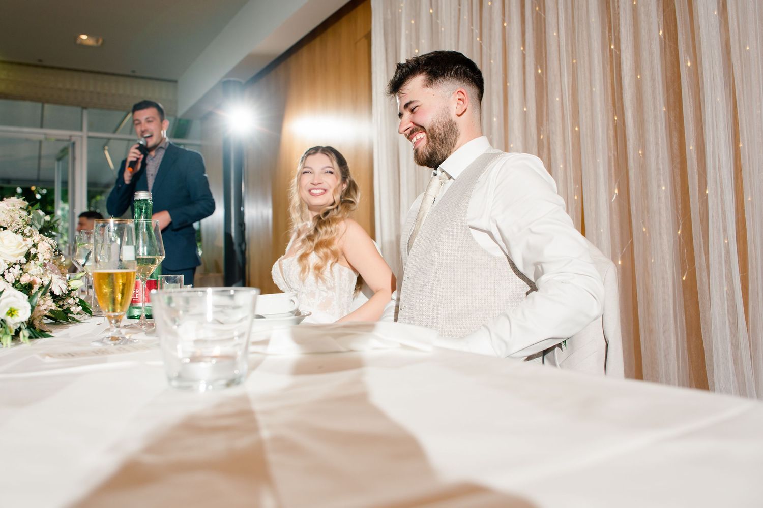 Bride and groom smiling at wedding reception table while guest delivers speech in background.