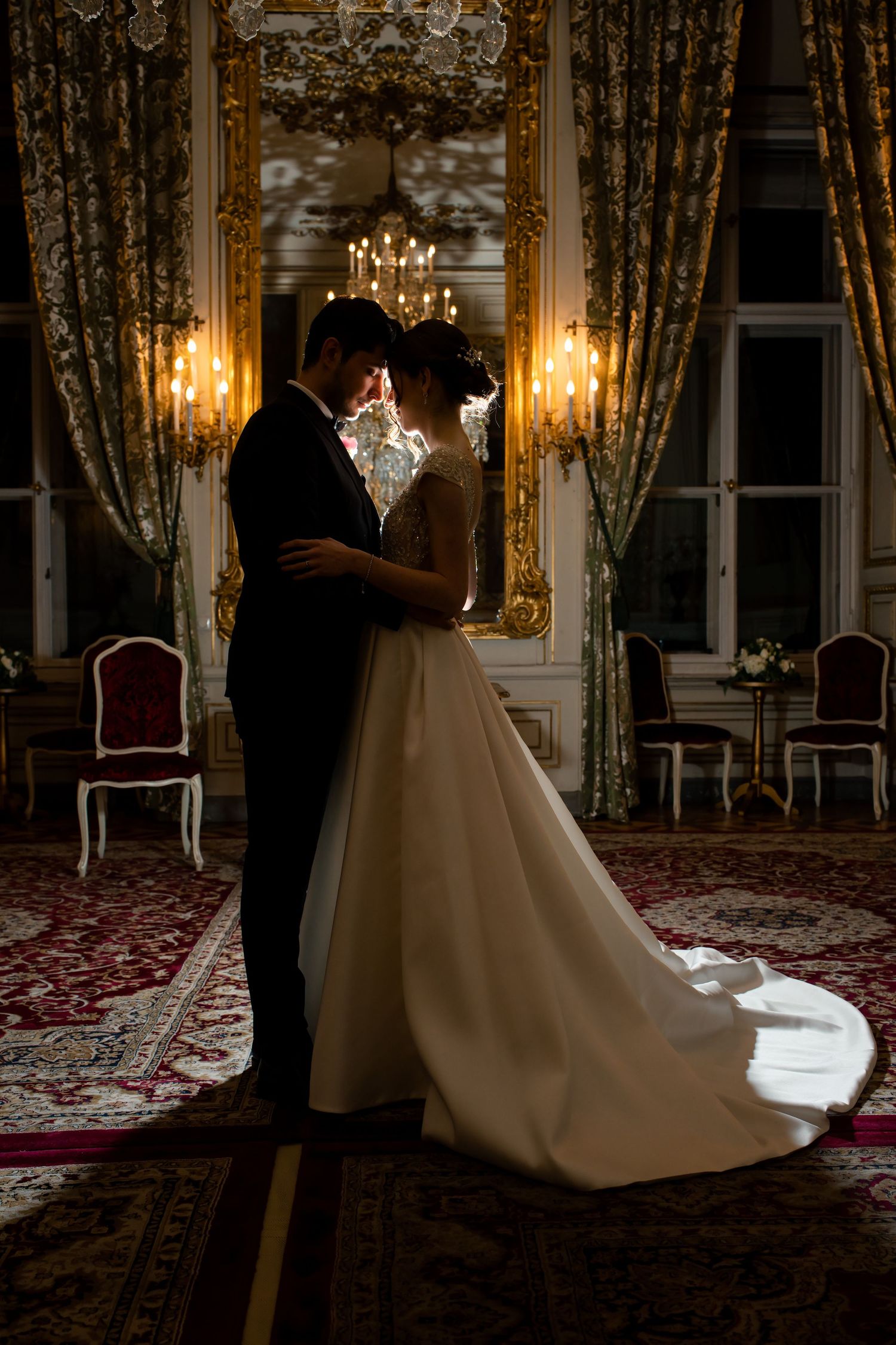 Elegant bride and groom embracing in a luxurious ballroom with golden chandeliers and ornate decor.