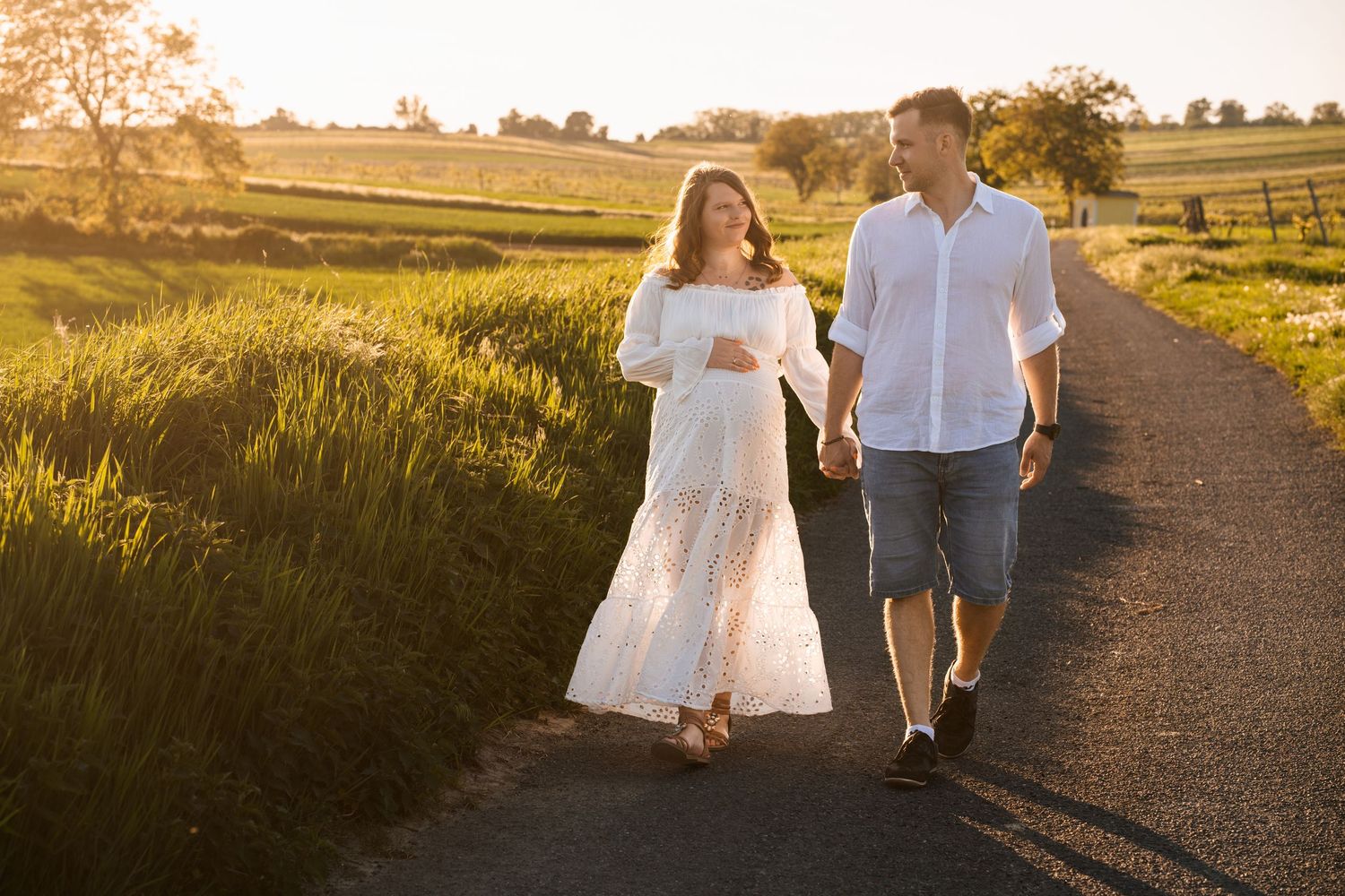A couple walks hand in hand down a sunlit path through a golden wheat field at sunset.