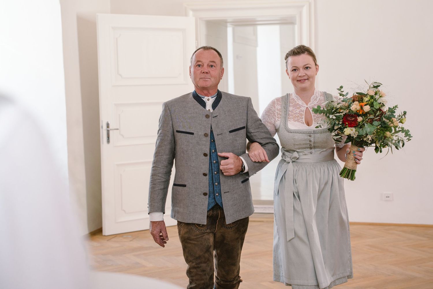 Two people in matching gray outfits stand together in a bright room with white walls and a potted plant.
