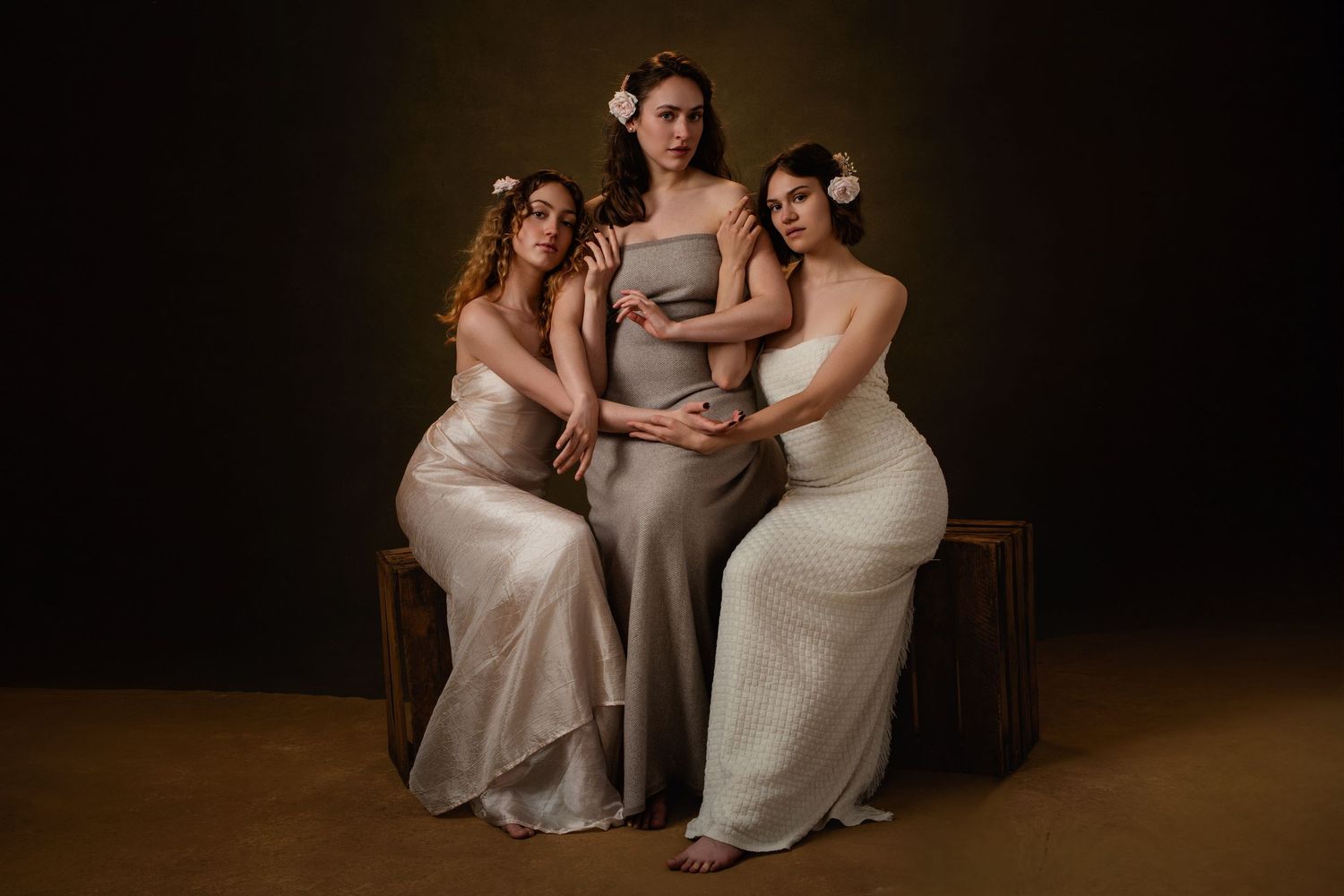 Women in neutral-toned dresses sit closely together against a dark background in an intimate portrait setting.