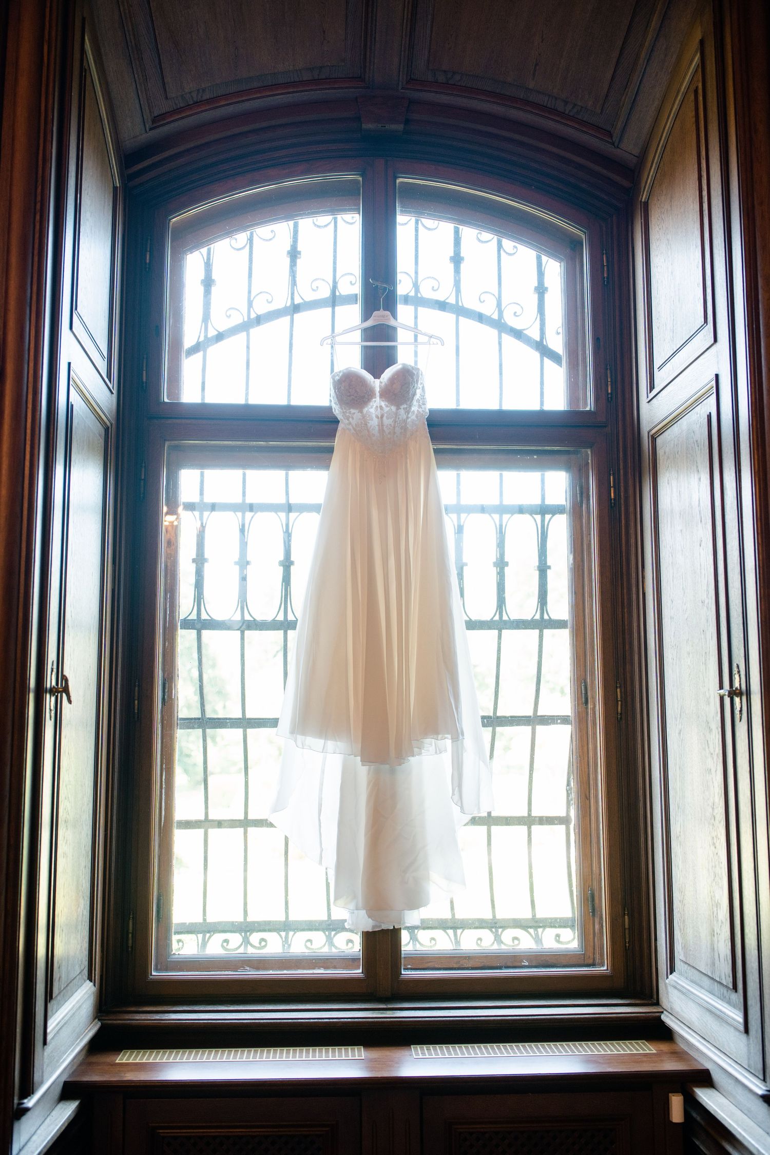 White wedding dress hangs elegantly in front of arched windows with ornate woodwork.