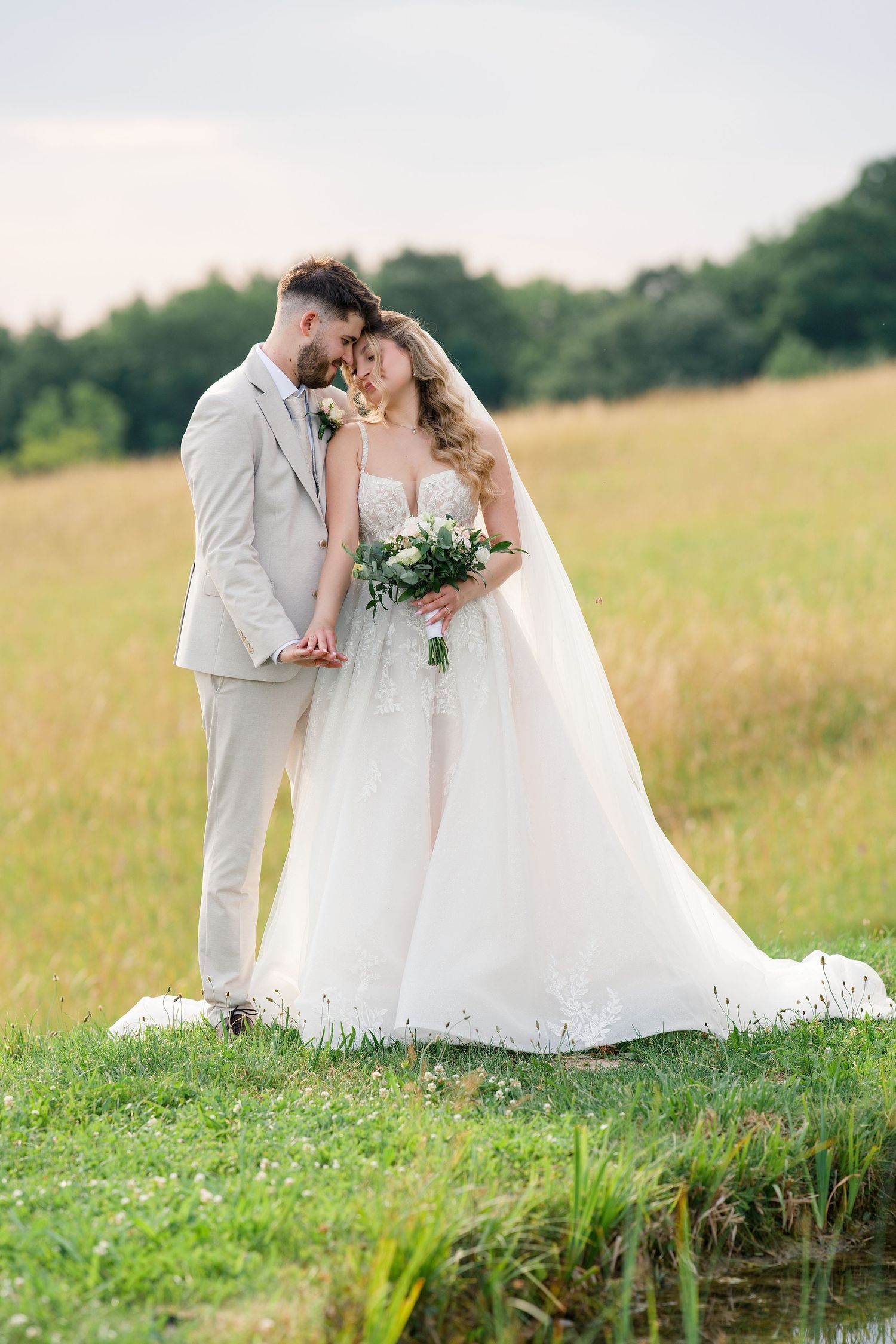 Bride and groom share a romantic moment in a sunlit meadow, bride holding a white bouquet in an elegant gown.