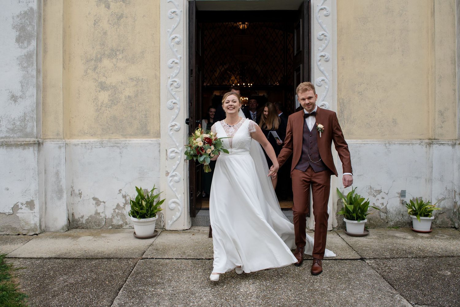 A couple exits a historic building entrance with cream colored walls and potted plants on their wedding day.