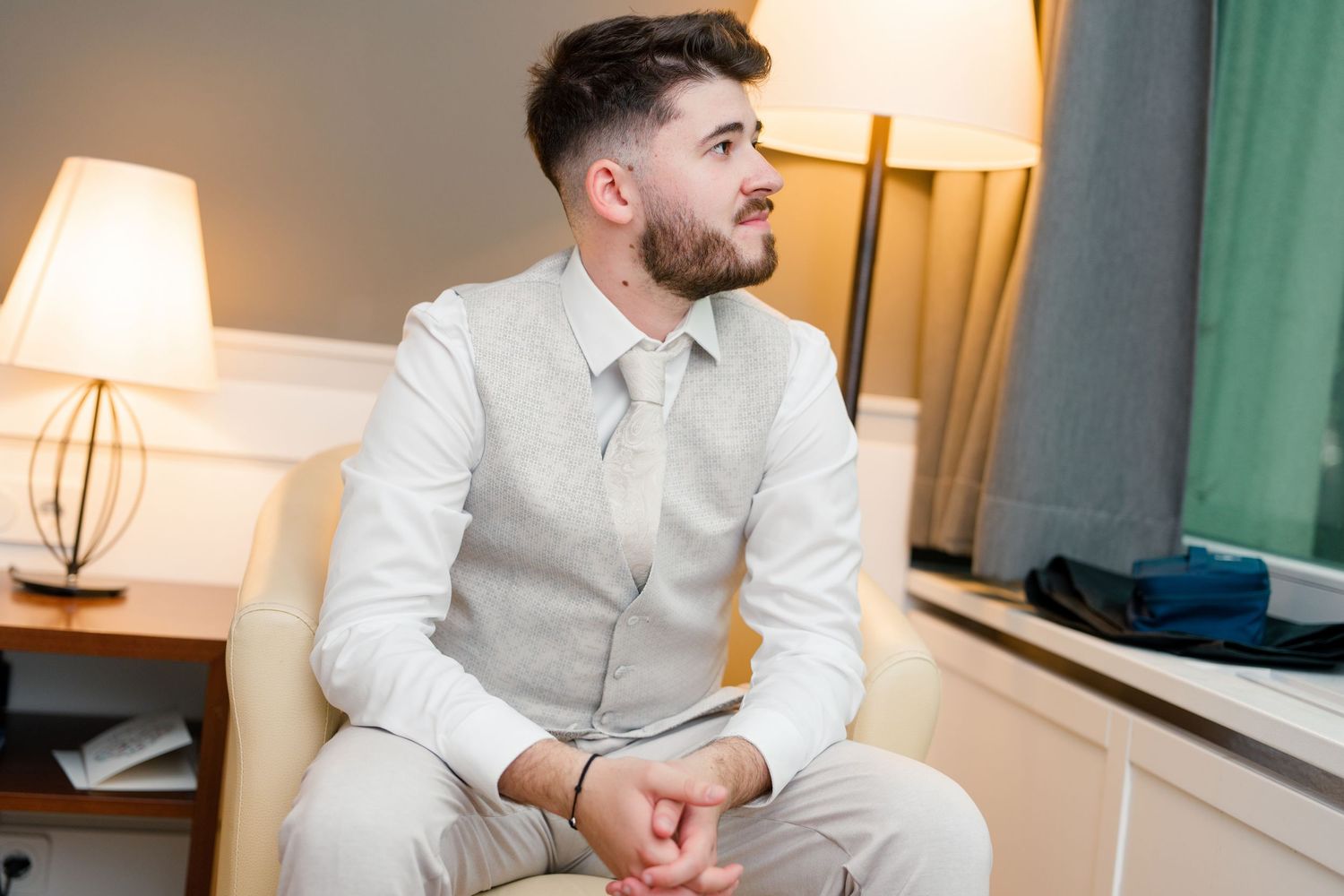 Elegant groom in white shirt and beige vest sitting in hotel room chair before wedding ceremony.