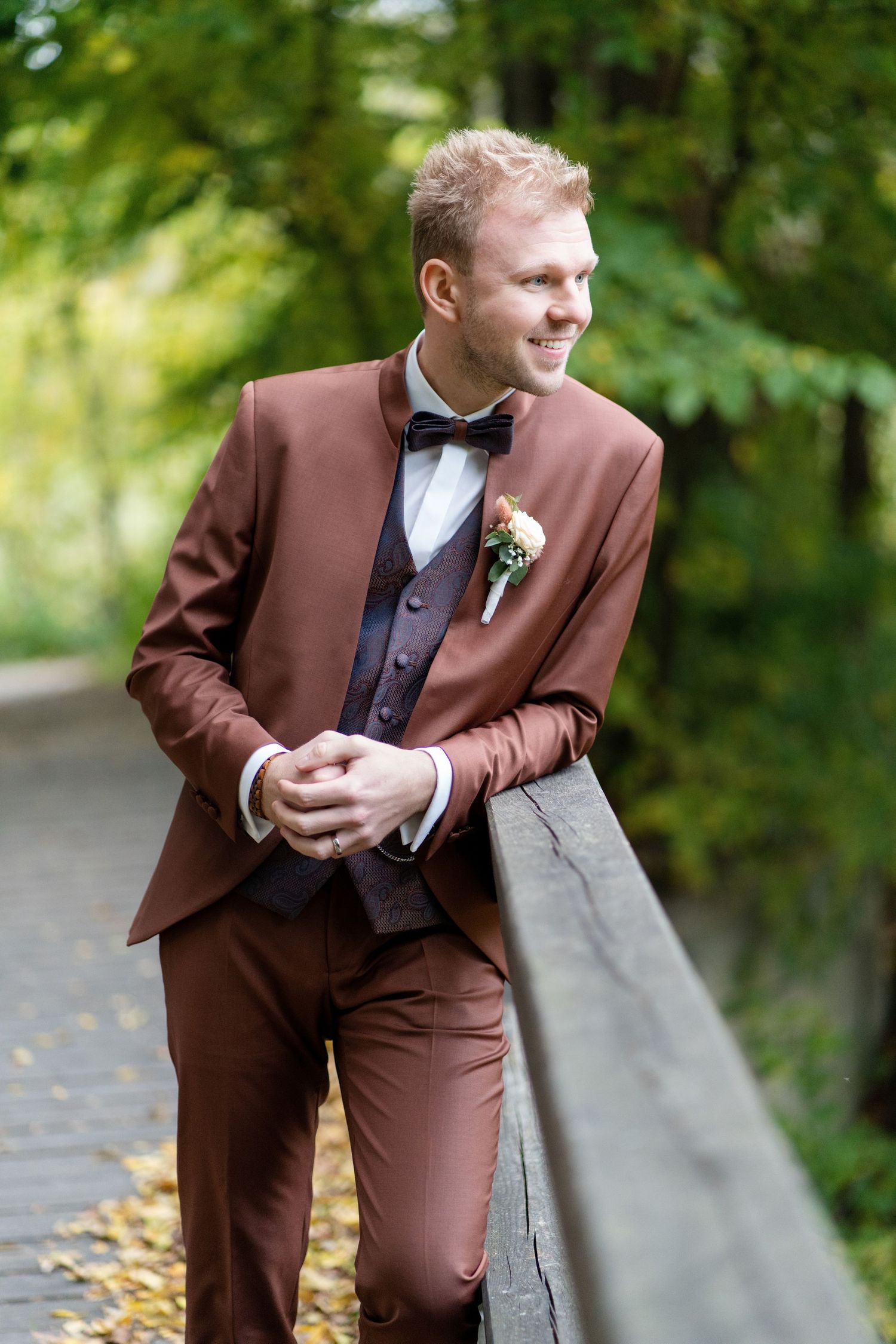 Groom in burgundy tuxedo poses playfully in outdoor garden setting.