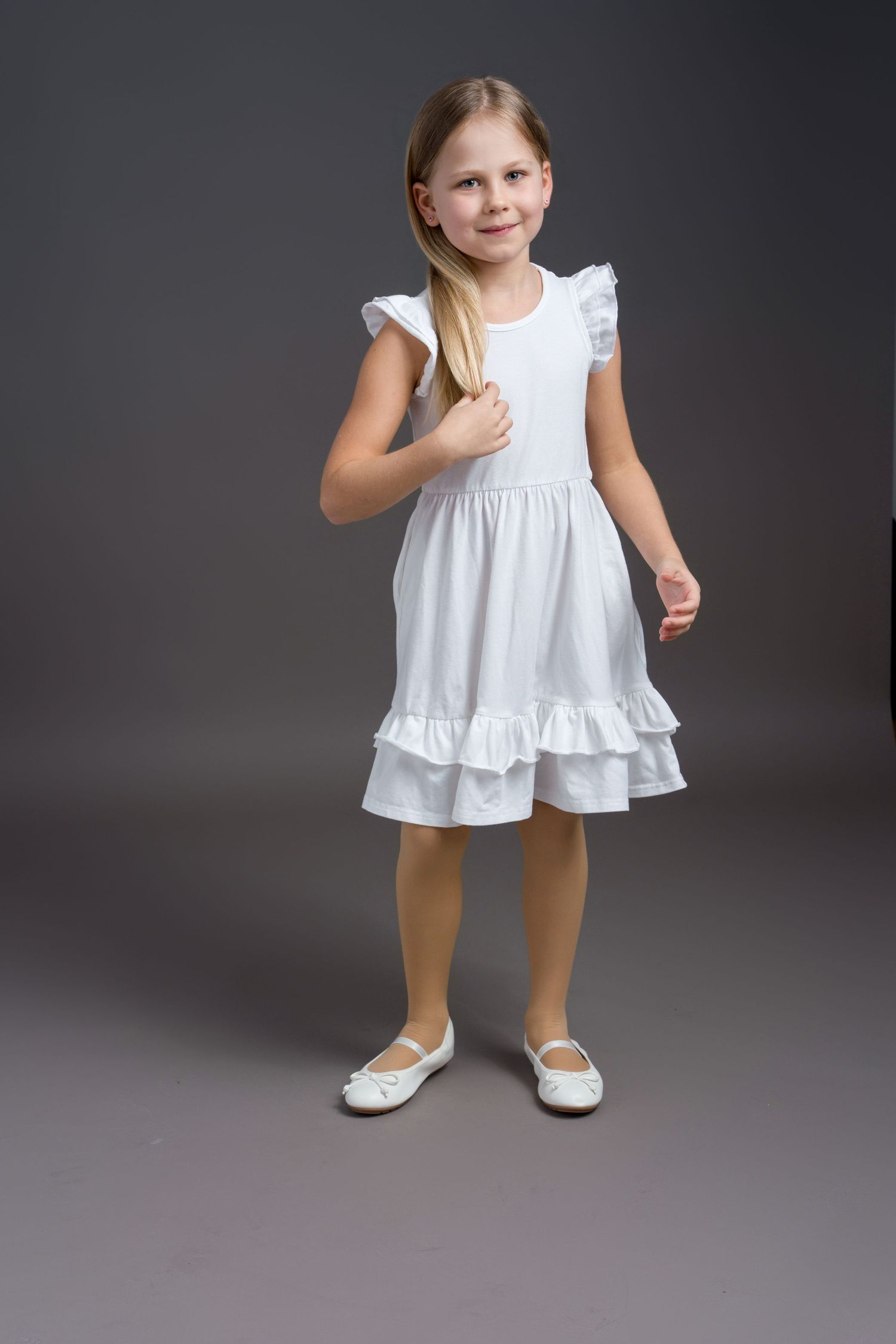 Young child in a white ruffled dress and white shoes poses against a dark gray studio backdrop.