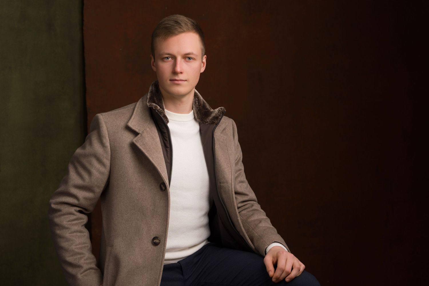 Young man in beige coat and white shirt posing confidently against a dark brown studio background.