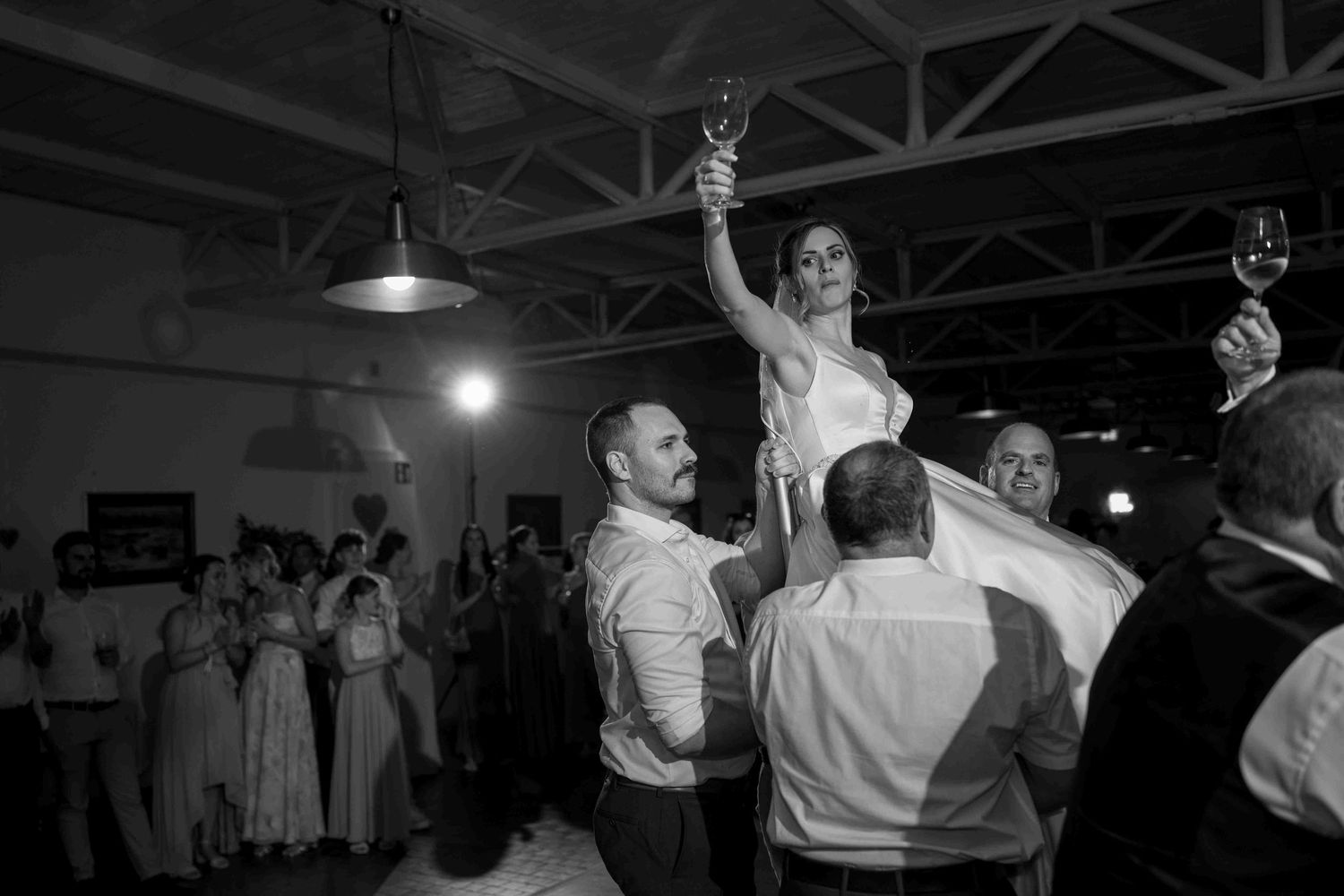 Black and white wedding photo of bride lifted by groom on dance floor, guests celebrating with champagne glasses raised.