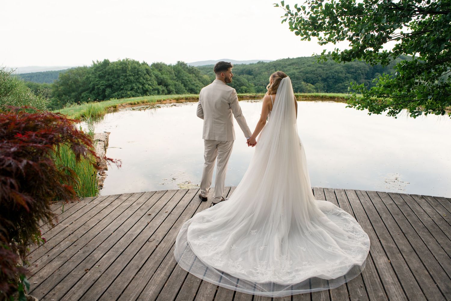 Bride and groom holding hands on wooden deck overlooking serene lake surrounded by lush green trees on wedding day.