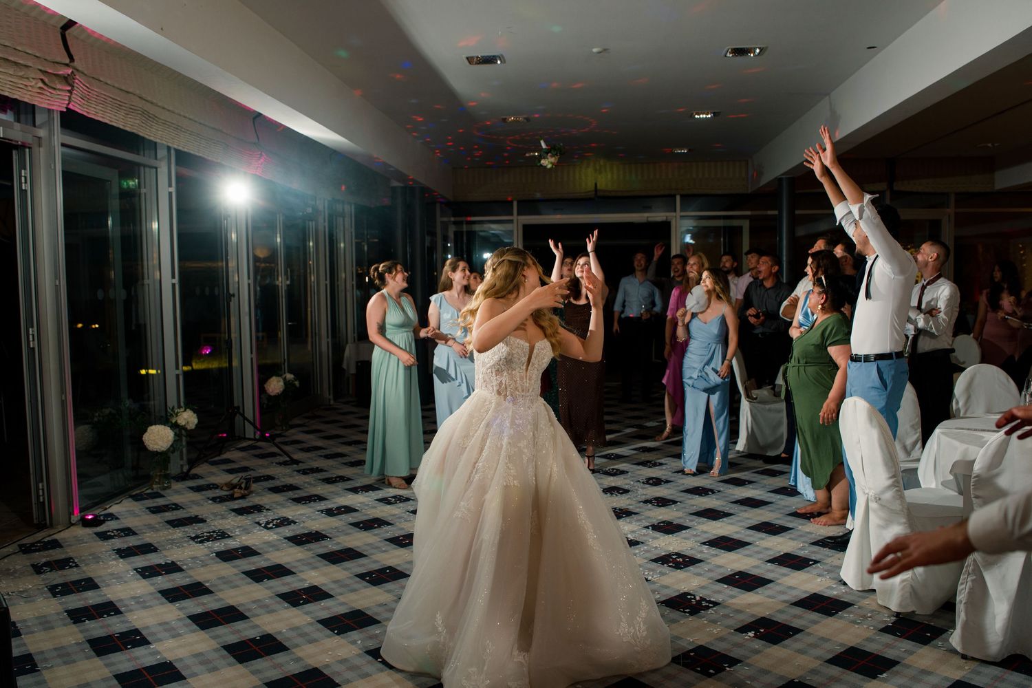 Bride in white ballgown shares first dance with guests celebrating on checkered dance floor at elegant wedding reception.