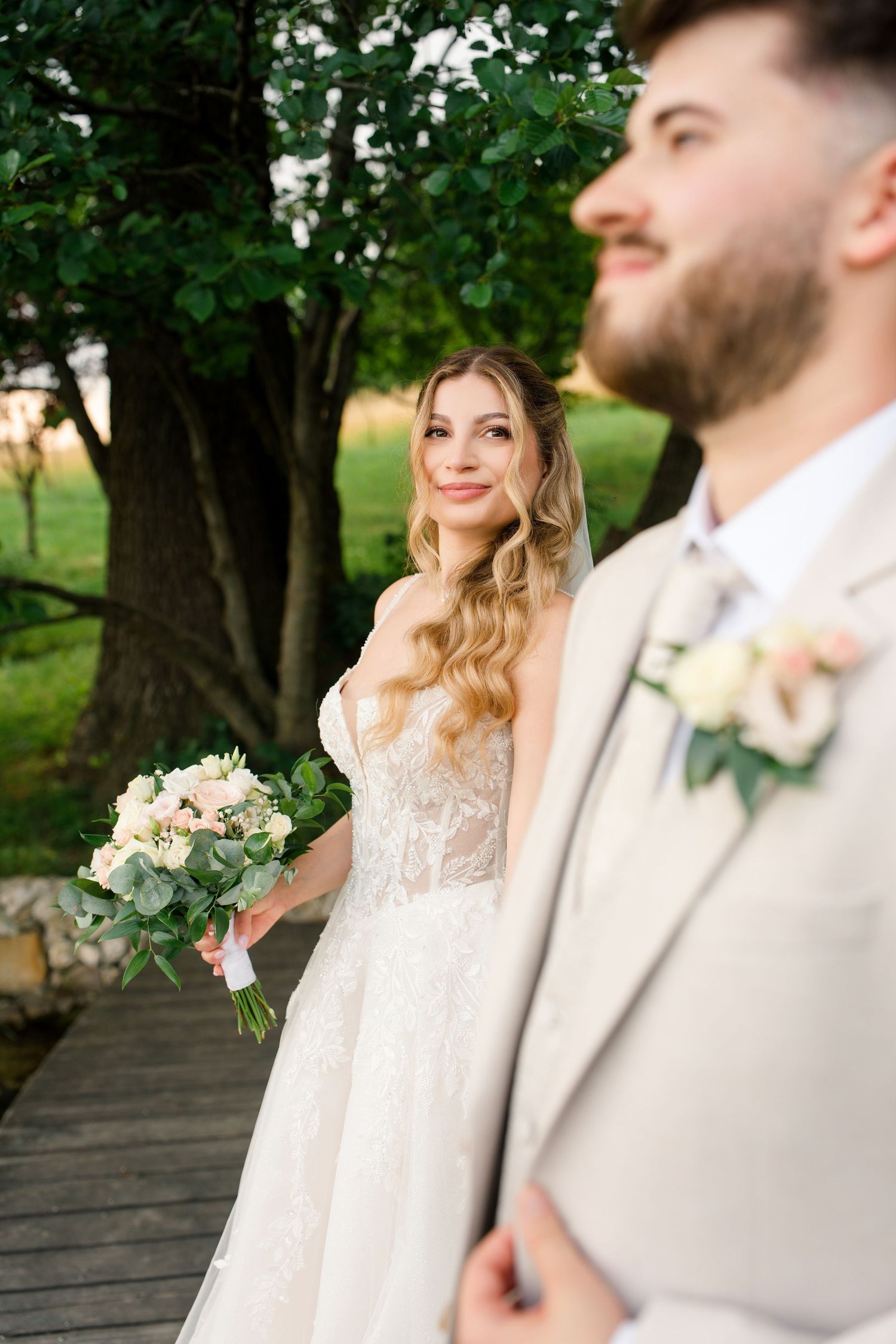 Smiling bride in lace gown holds bouquet beside bearded groom in beige suit outdoors near green trees.
