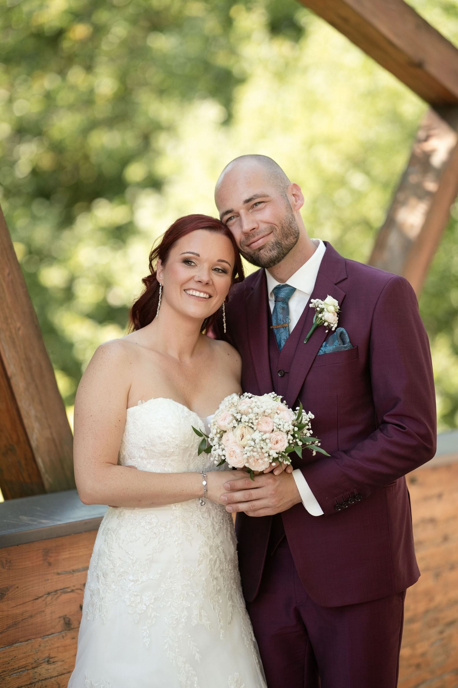Bride in white lace gown and groom in burgundy suit pose together holding a bouquet outdoors at their wedding.